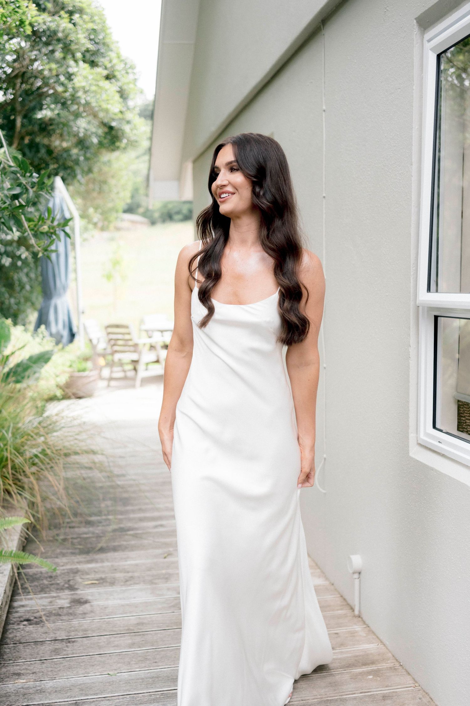 Woman in long white silk dress stands on wooden deck by house exterior.