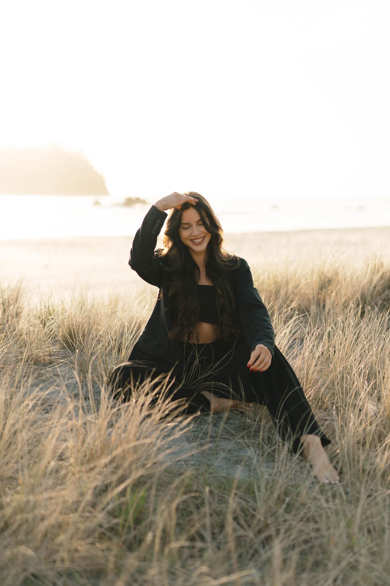 Person in black outfit sits playfully in tall grass at sunset.