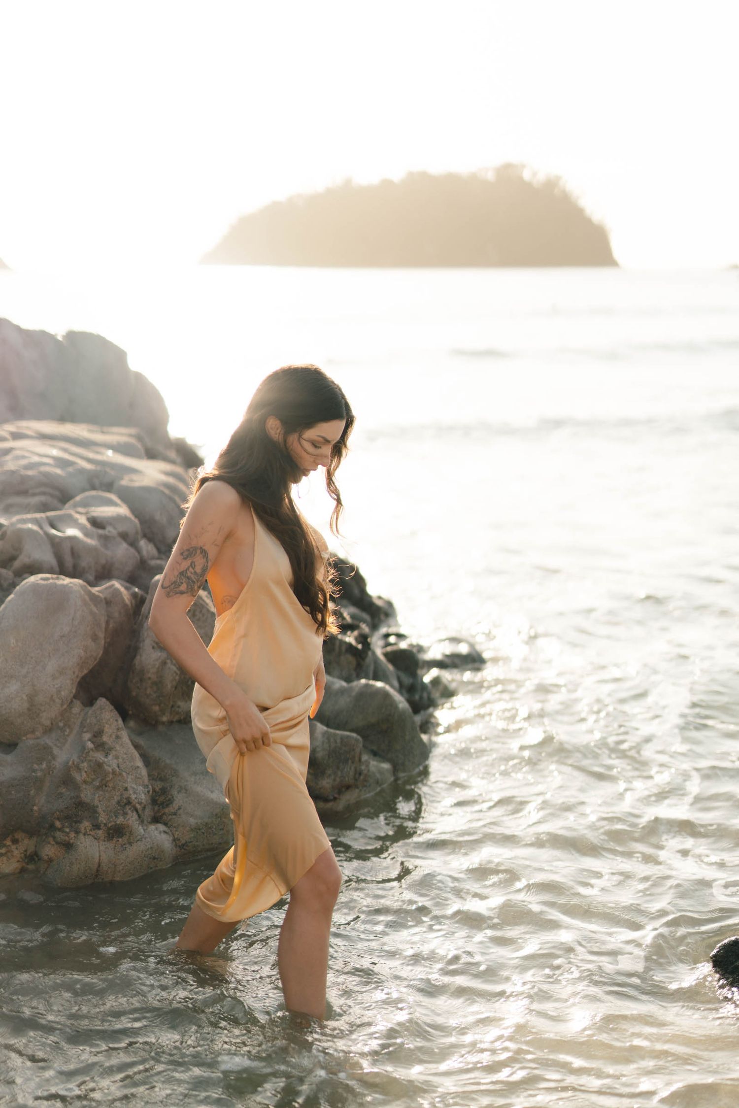 Person in a yellow dress posing gracefully among coastal rocks with ocean waves behind.