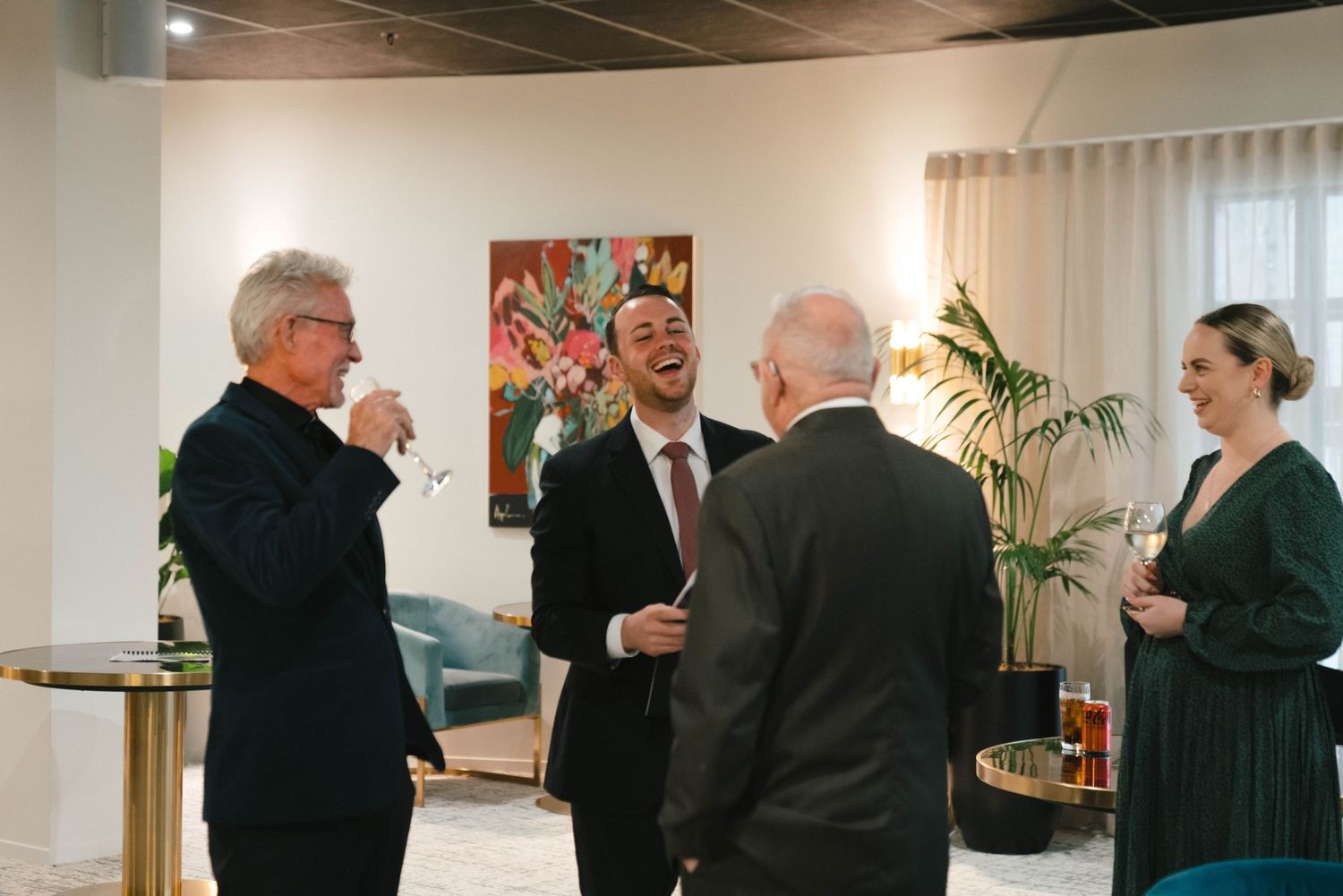 A group of people conversing at a social gathering in a modern office space with a colorful painting on the wall.