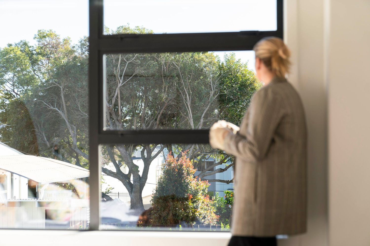 Person in gray sweater looking out large window at suburban landscape.