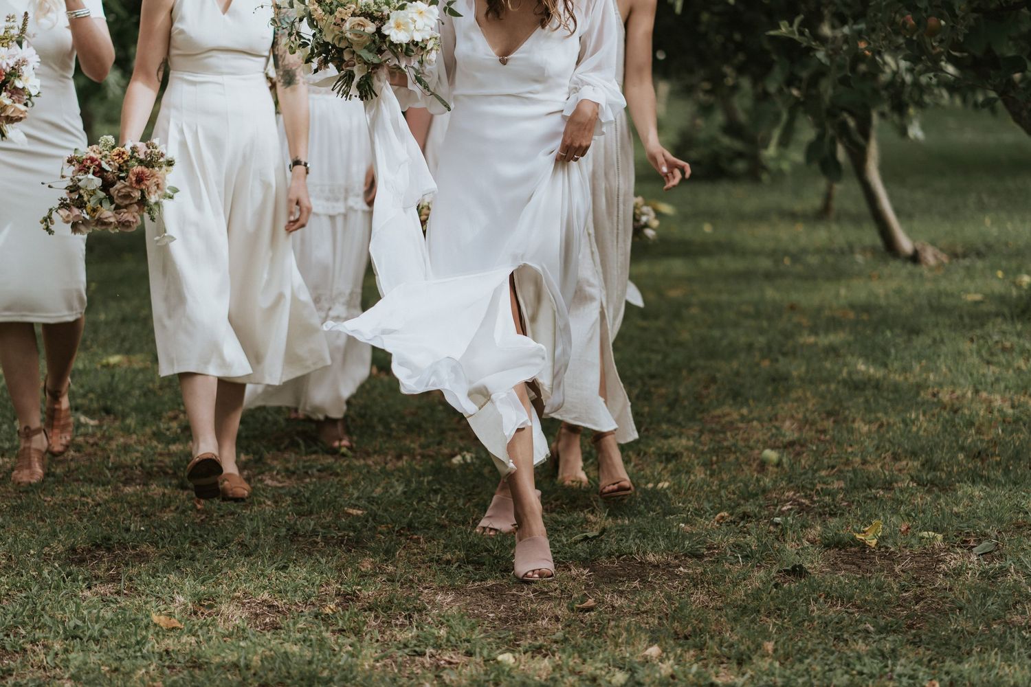 Multiple bridesmaids in flowing white dresses walk barefoot through grass while holding floral bouquets.