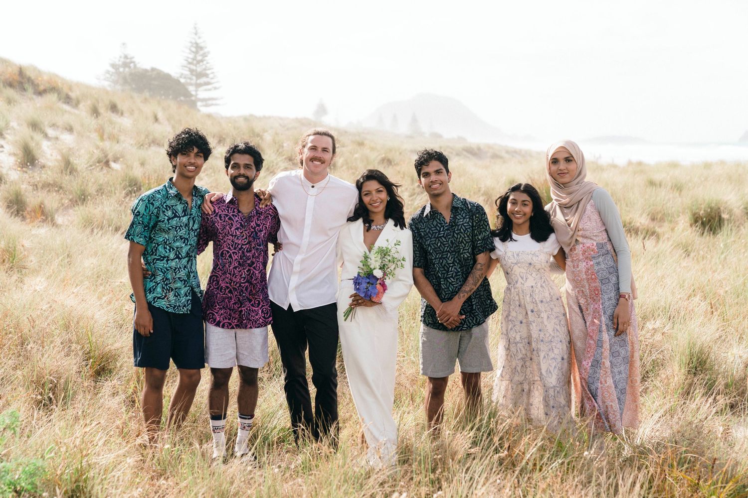 Group photo of wedding party in casual attire standing together on grassy hillside.