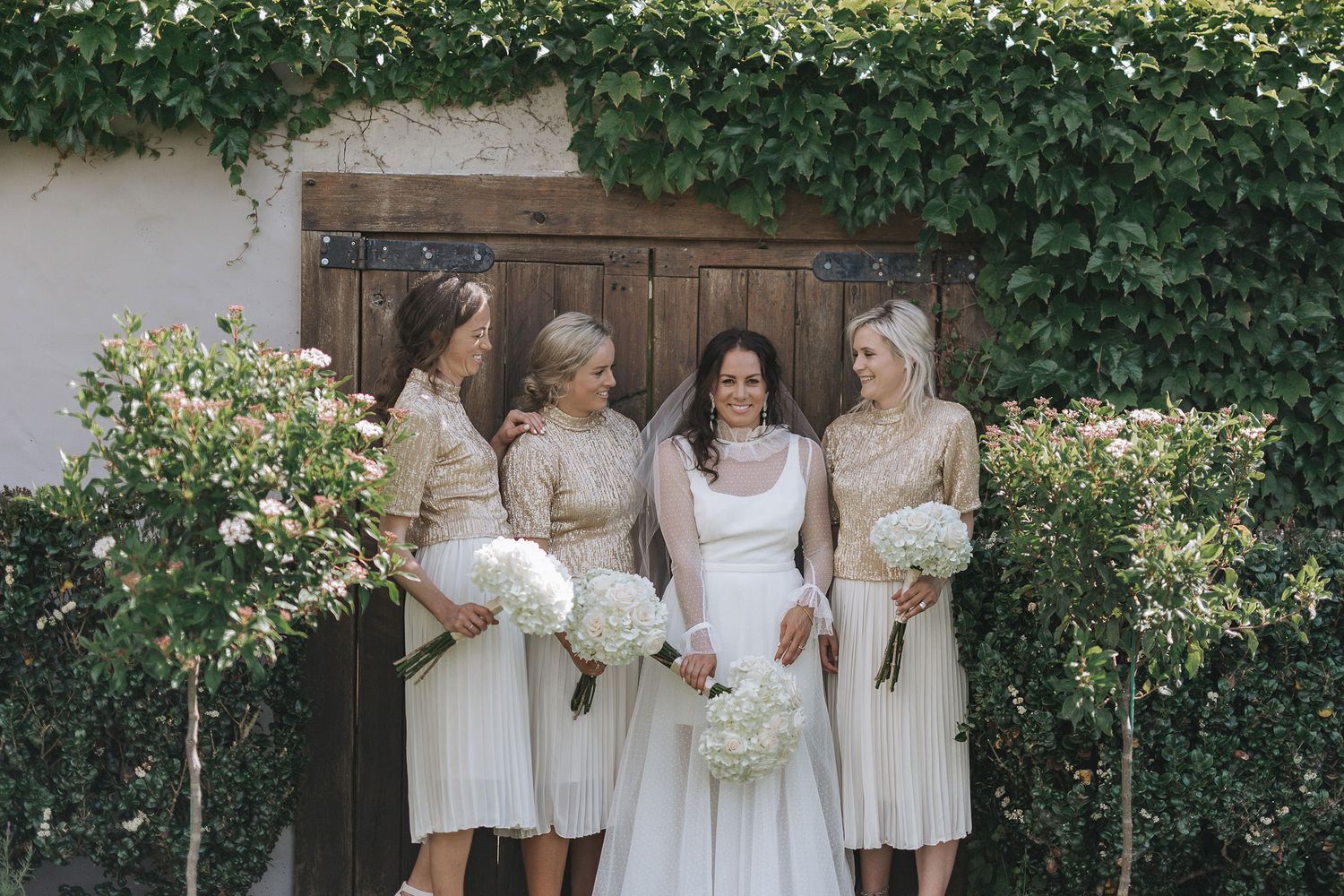 A wedding party in light cream and white dresses poses together against a rustic backdrop with climbing vines.