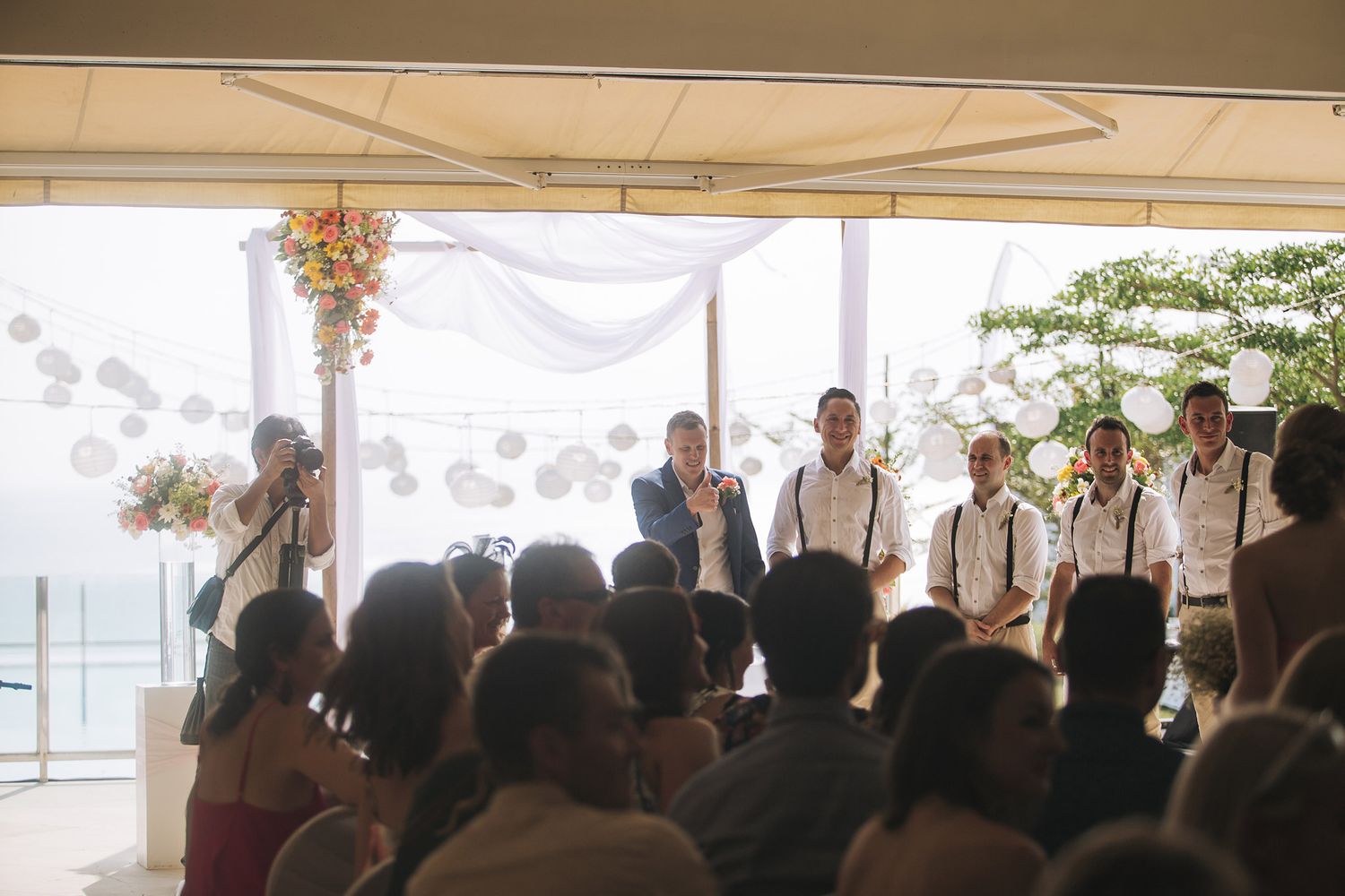 Outdoor wedding ceremony with guests under white tent by water.