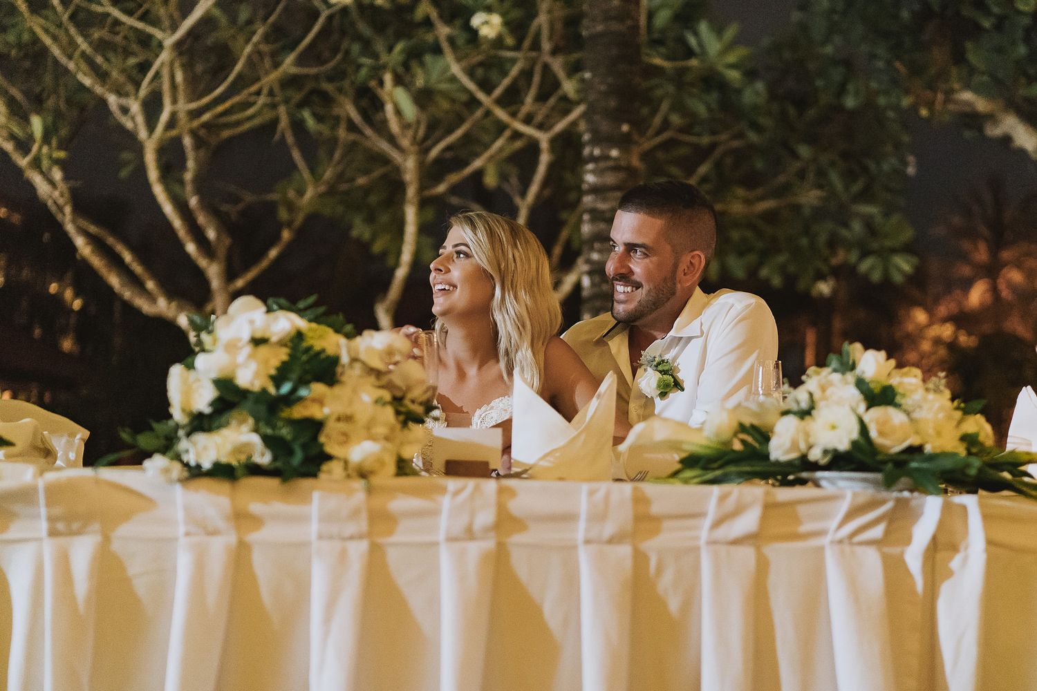 A newlywed couple sits at their elegant white-draped wedding reception table decorated with yellow and white flowers.