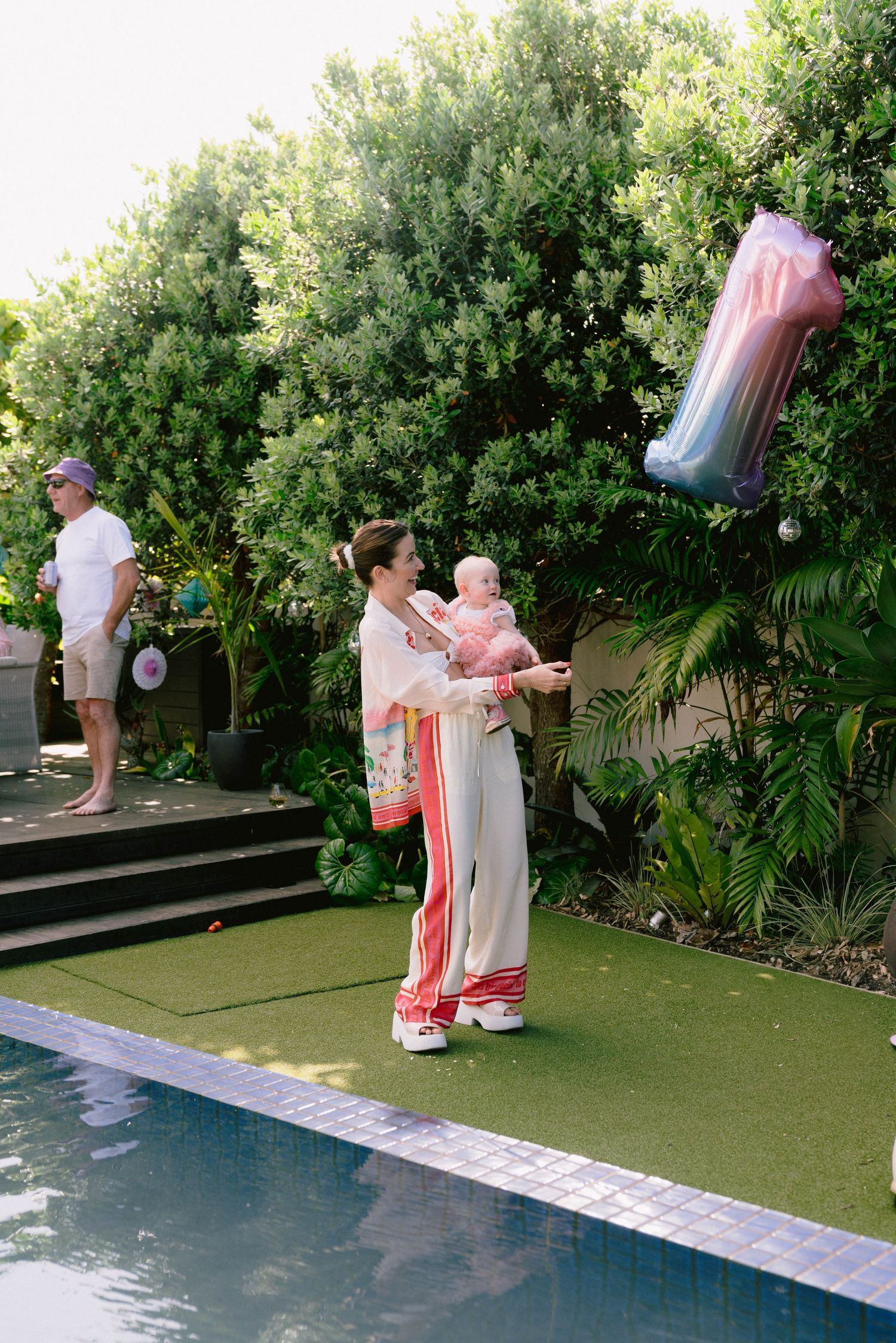 Guests celebrate at an outdoor pool party with pink decorations and lush greenery in the background.