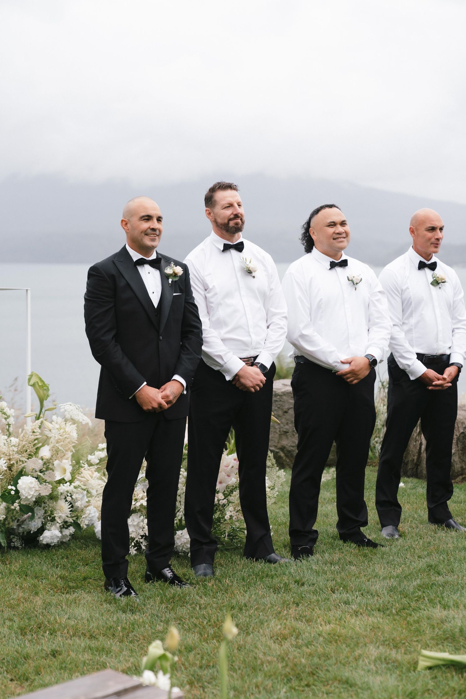 Groomsmen in formal black and white attire stand together at outdoor ceremony with water view.
