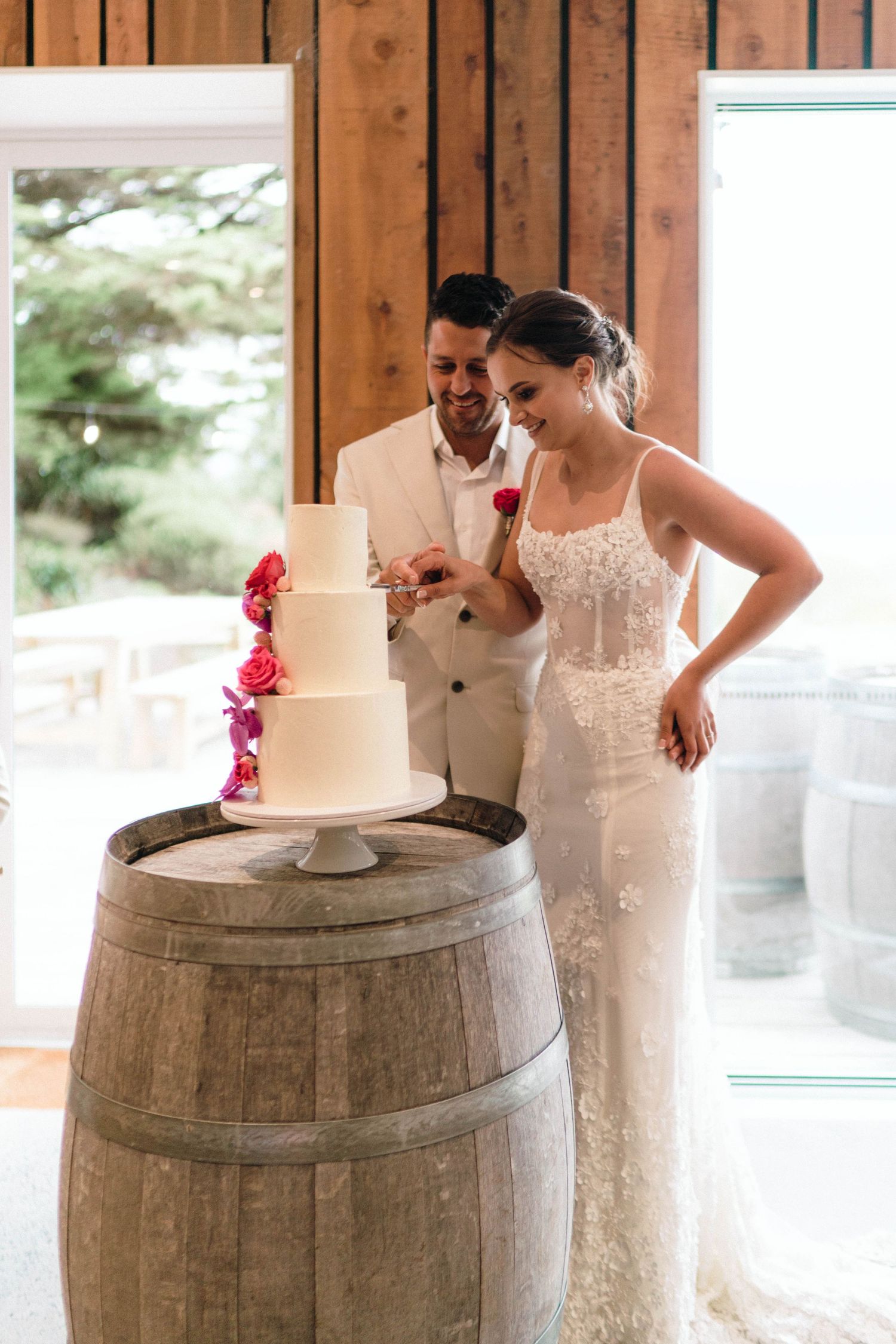 Three-tiered white wedding cake with pink flowers displayed on rustic wine barrel.