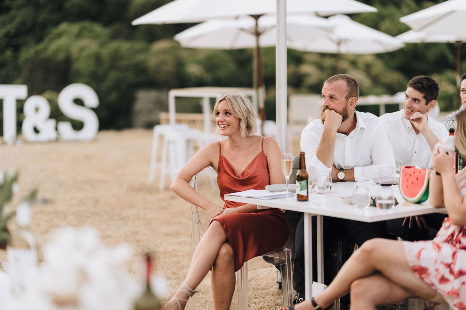 Wedding guests seated at outdoor tables with white umbrellas enjoy the reception festivities.