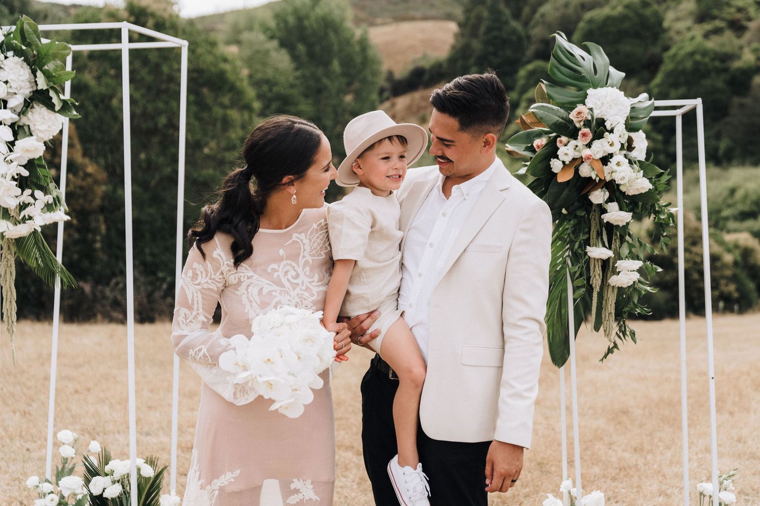 A couple in white formal attire holds a small child during their outdoor wedding ceremony.
