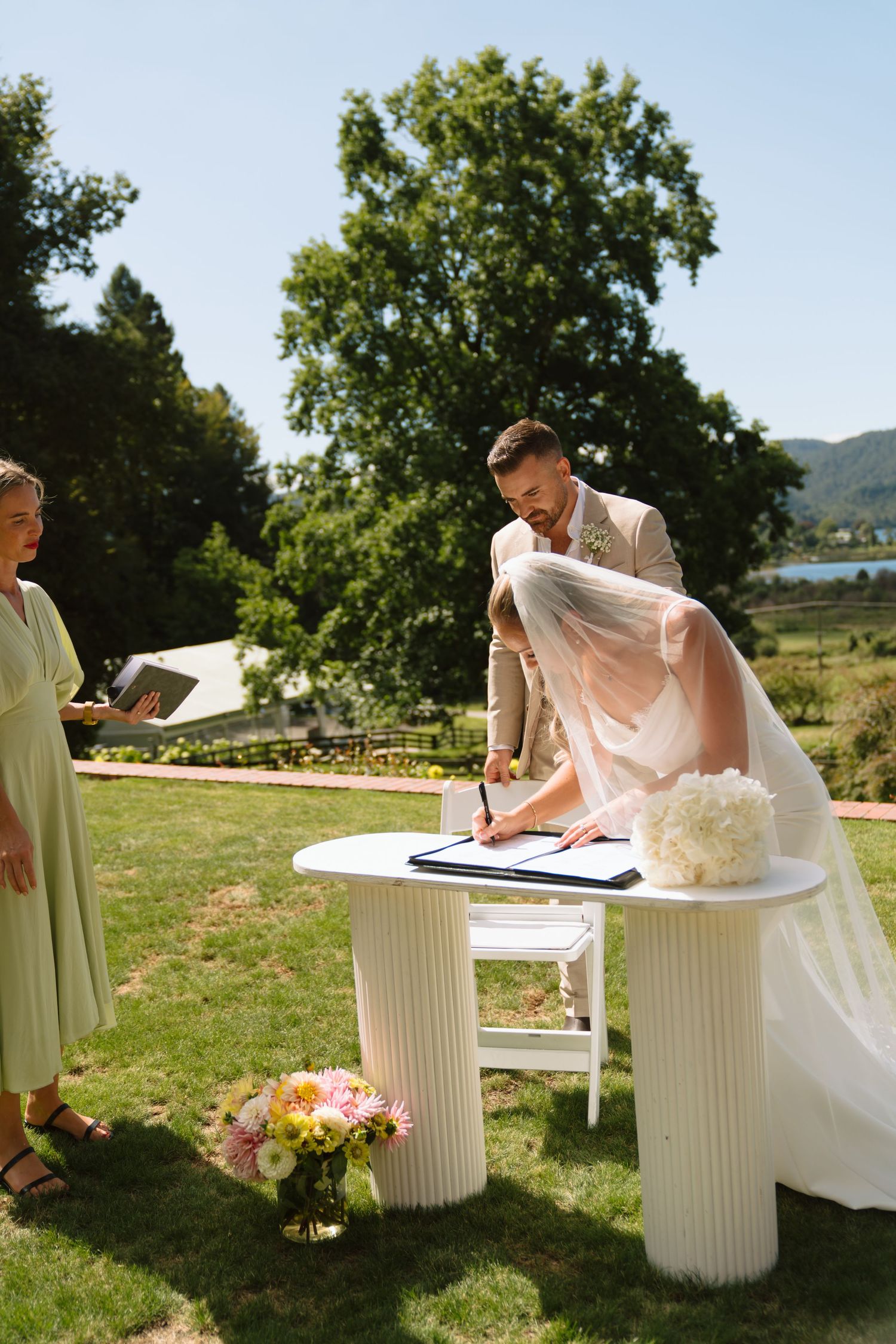 Wedding guests sign documents on white columns outdoors in a sunny garden setting with mountains in background.