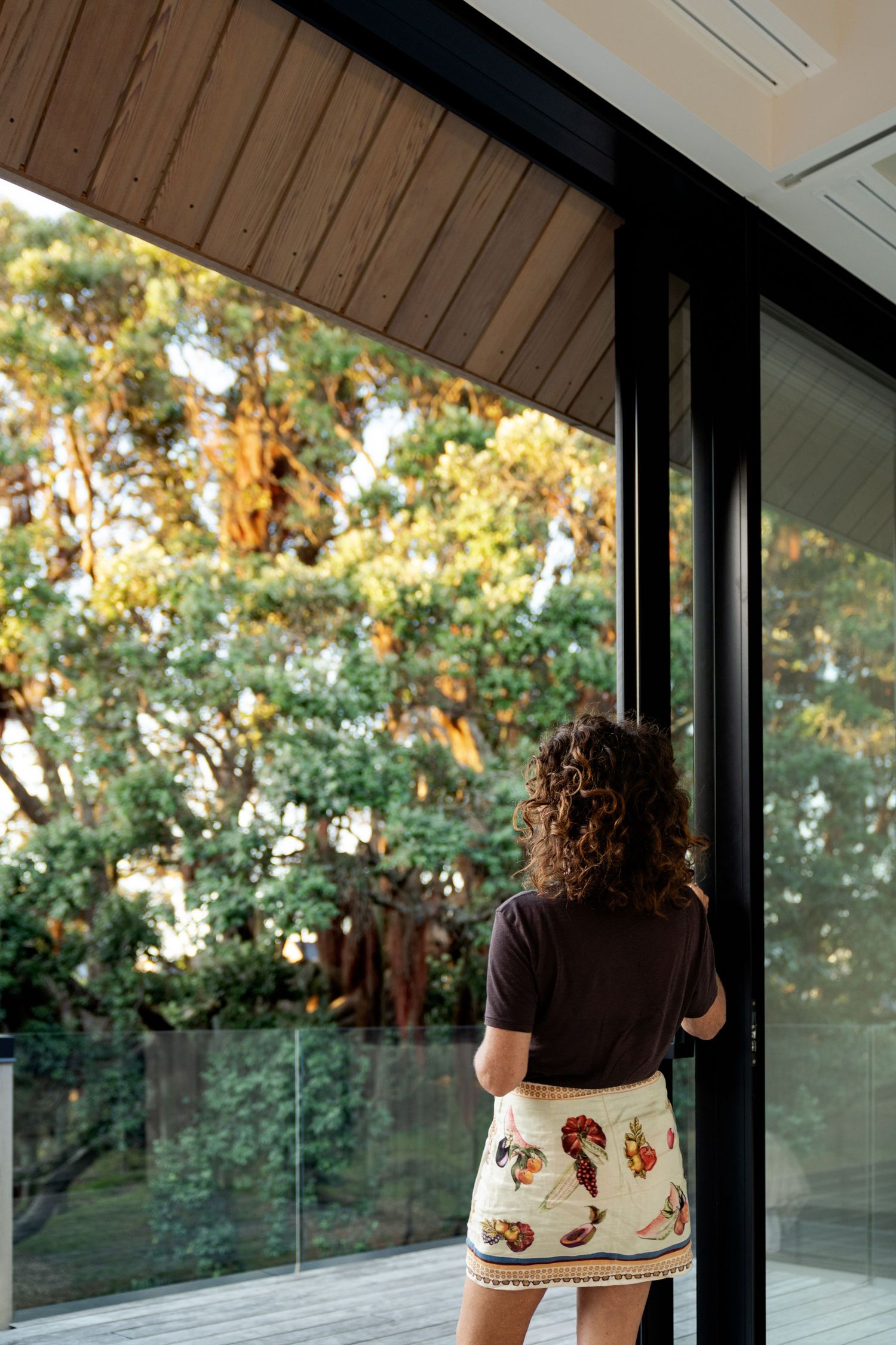 Person wearing a black top and patterned skirt stands on a deck with glass railing and trees in the background.
