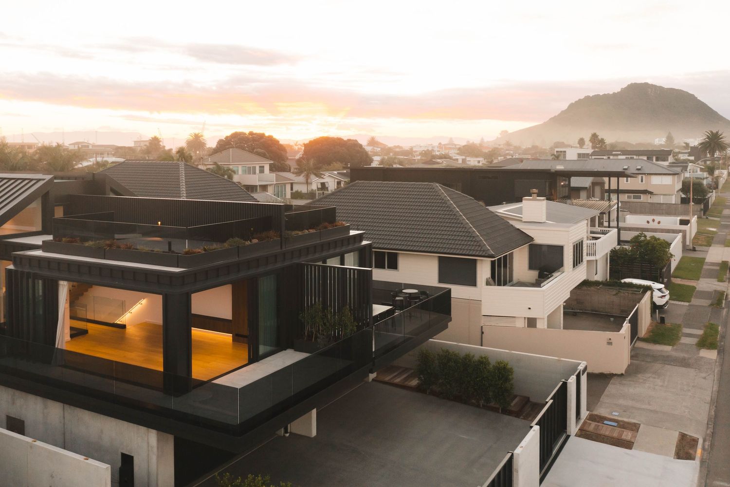 Contemporary black beach houses with illuminated windows at sunset against mountain backdrop.
