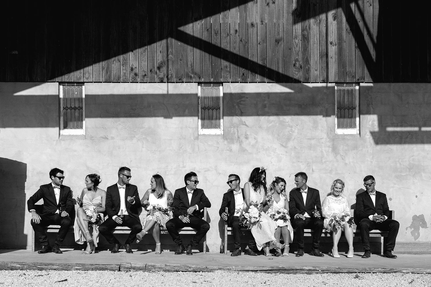 Large panoramic black and white photograph showing a row of people seated on a bench against a wall.
