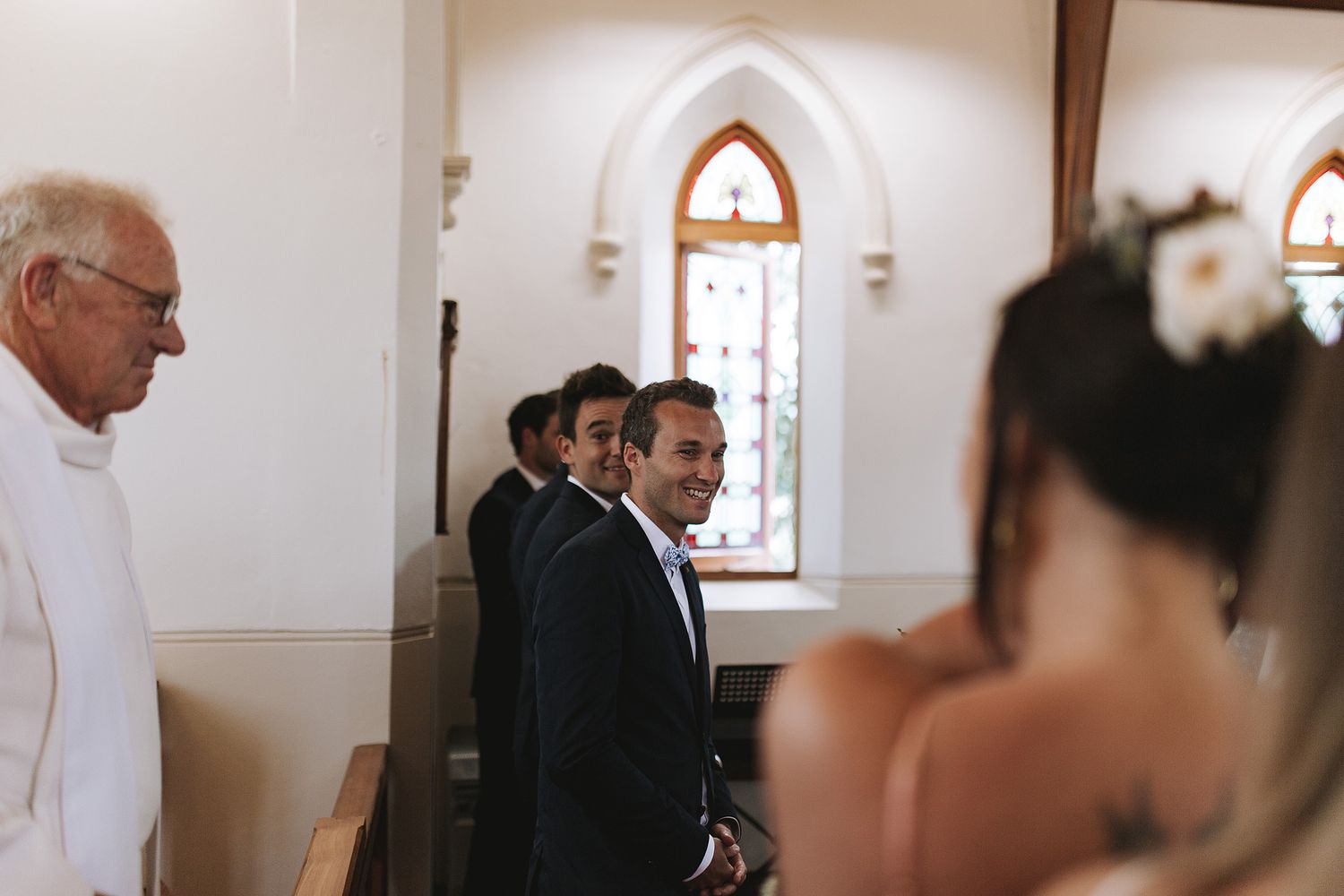 Wedding guests are gathered in a bright church sanctuary with stained glass windows and arched doorways.