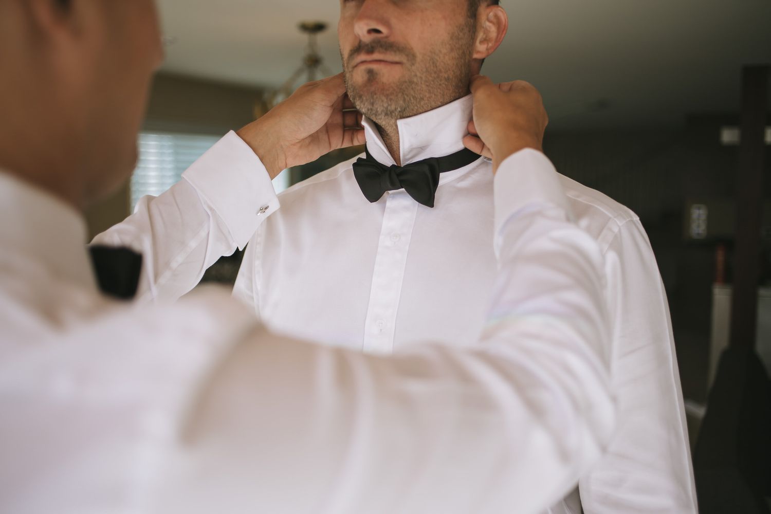 Someone helps adjust a black bow tie on a groom wearing a crisp white dress shirt during wedding preparations.
