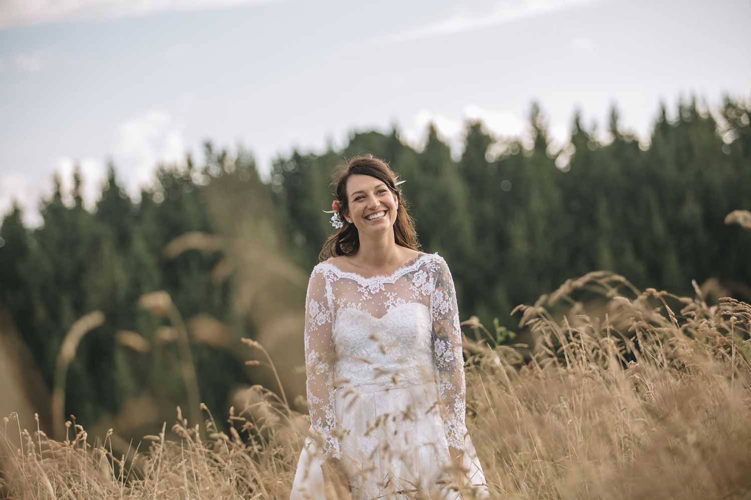 A person in a white lace wedding dress stands joyfully in a golden wheat field with evergreen trees in the background.