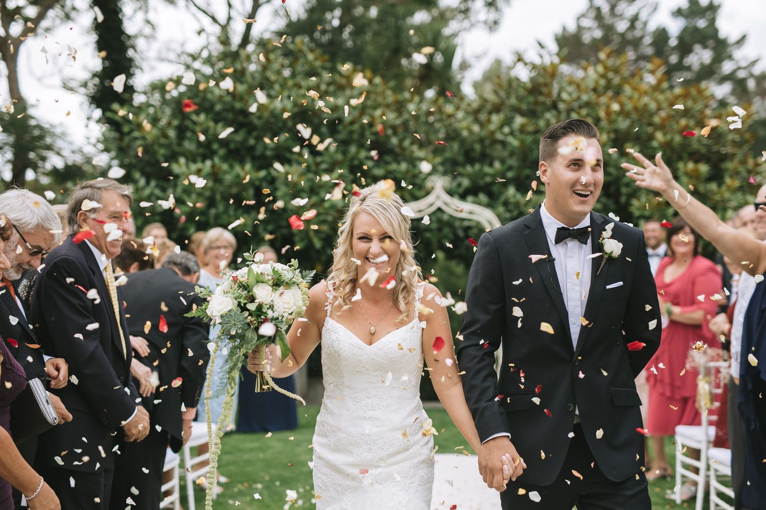 A couple walks through a shower of confetti on a garden lawn during their wedding celebration.