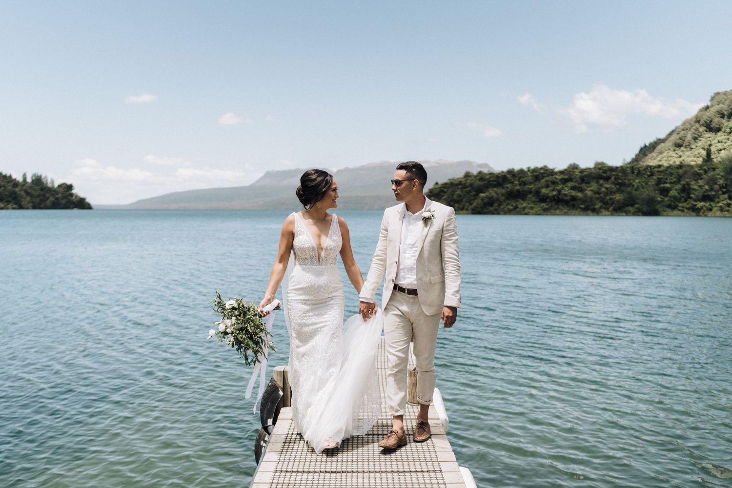 Newlyweds stand on a dock overlooking a serene lake surrounded by mountains on their wedding day.