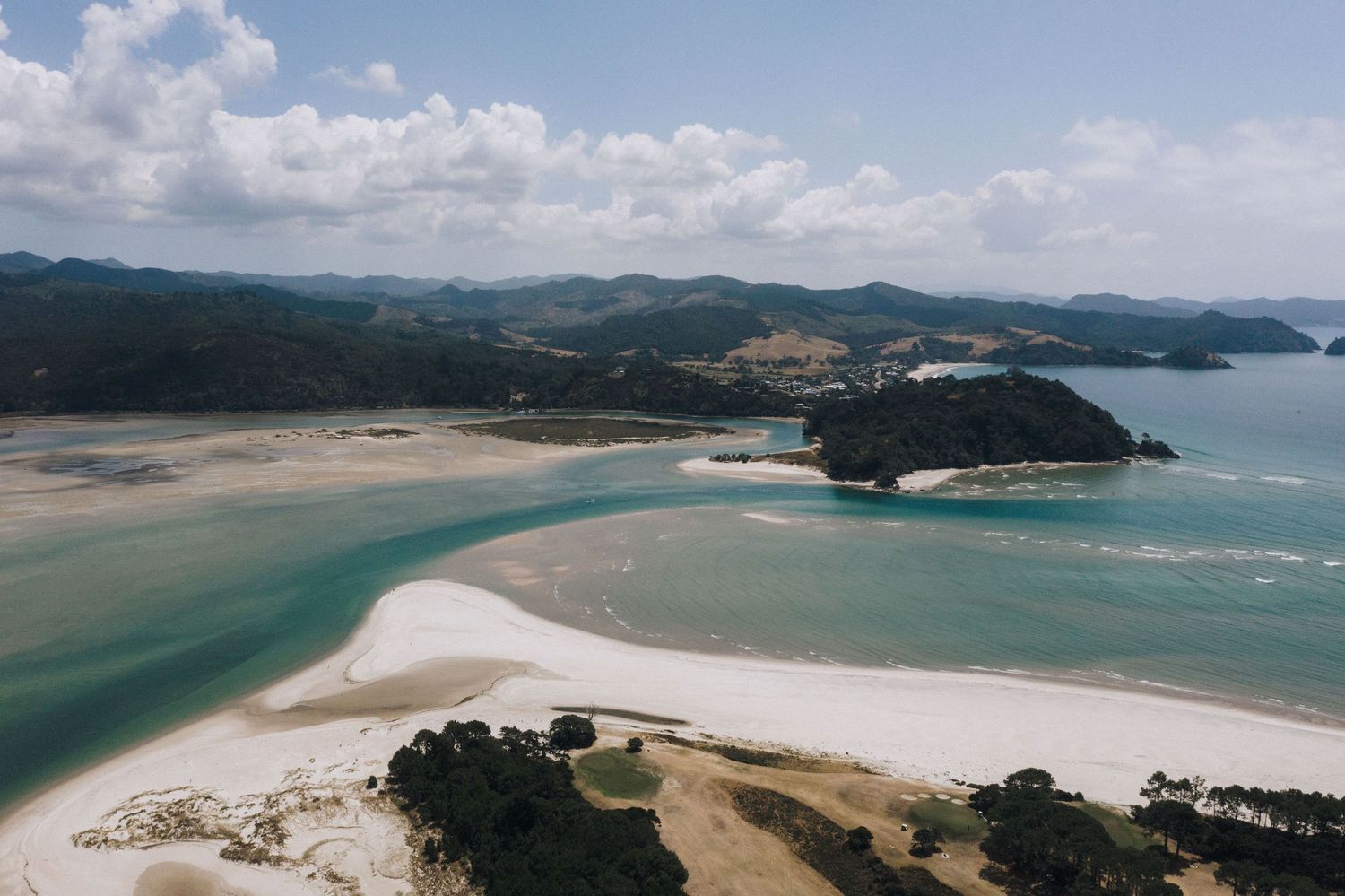 Aerial view of turquoise waters and white sand formations along a coastal landscape with forested hills and islands.