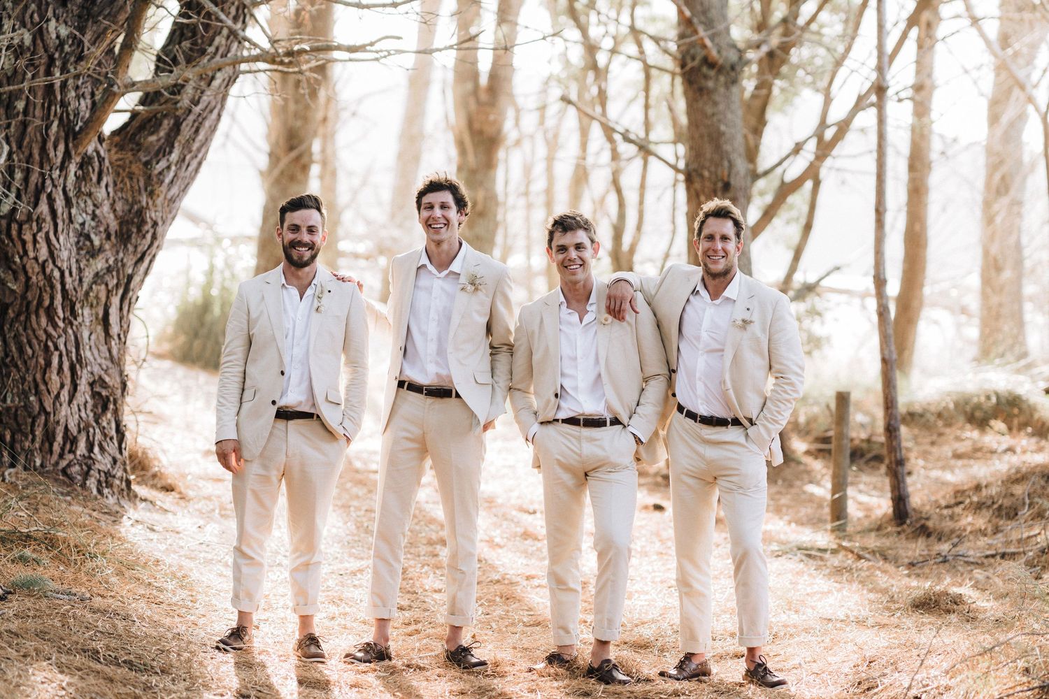 Four groomsmen in matching white shirts and khaki pants pose together in a sunlit forest.