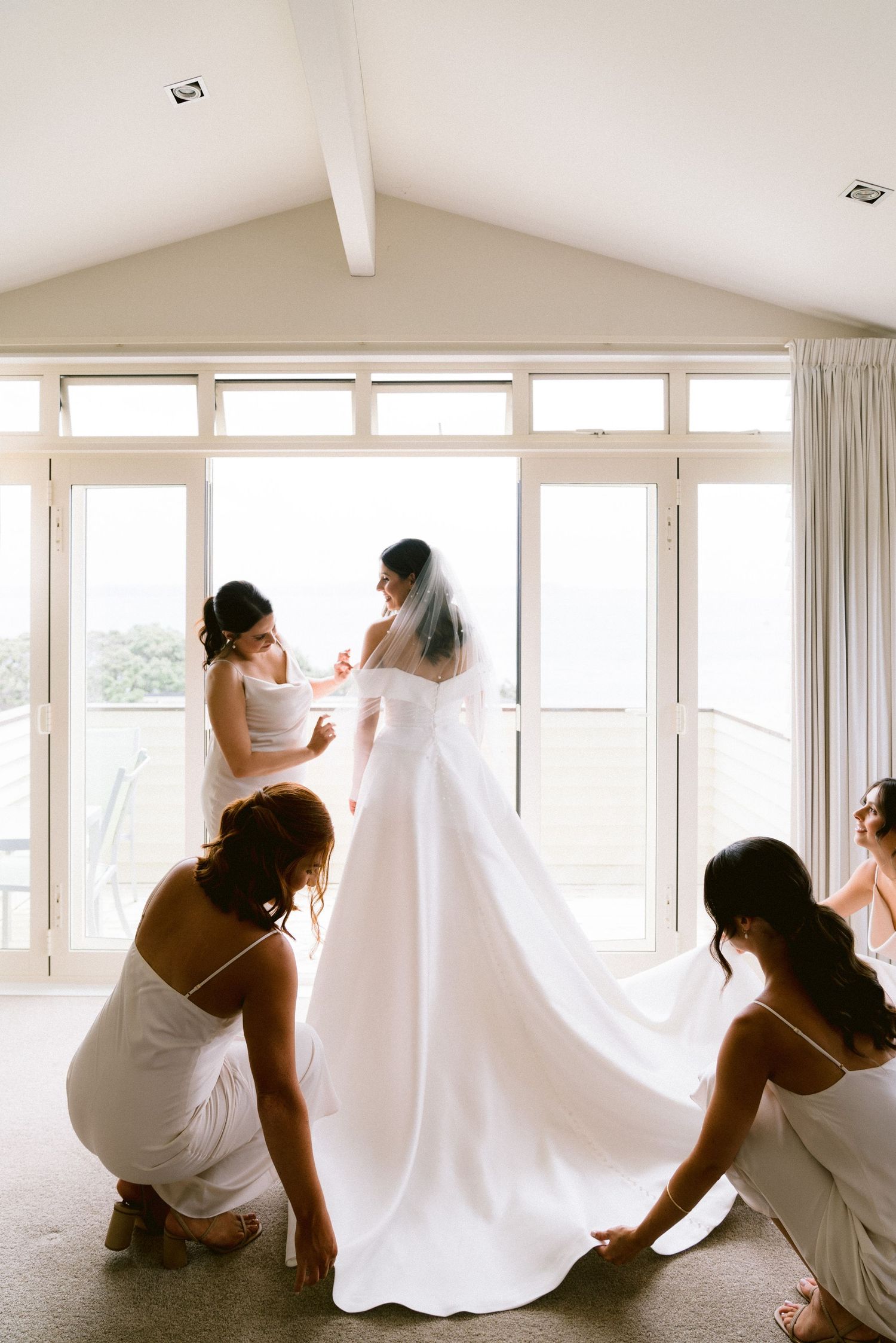Bridesmaids in white dresses gather at a window-filled room creating candid emotional moments together.
