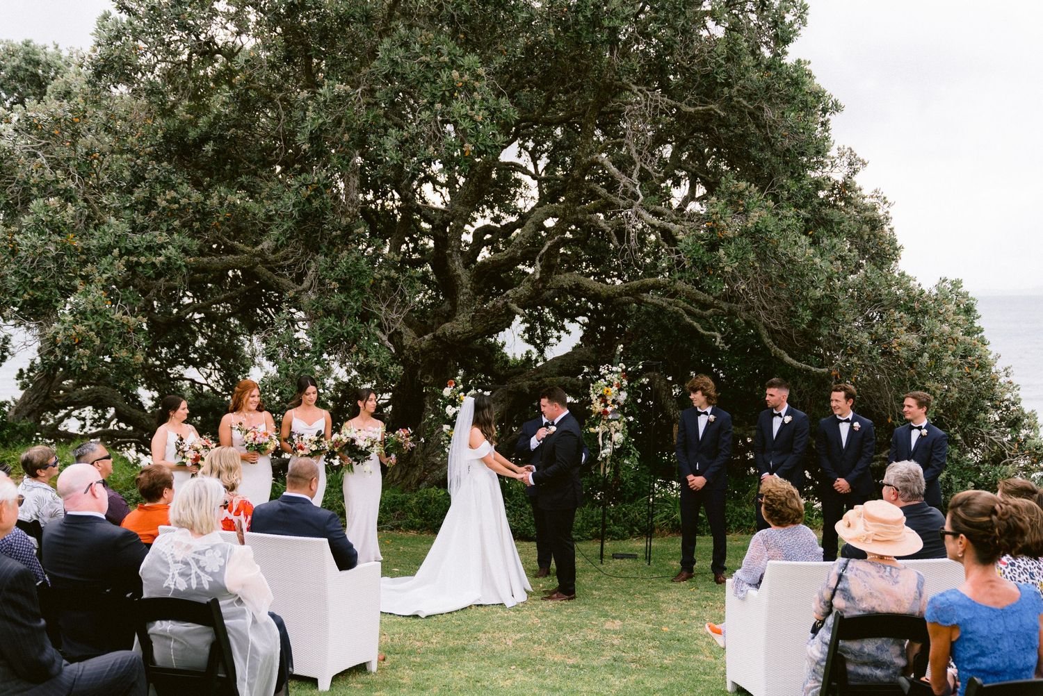 Outdoor garden wedding ceremony under a majestic oak tree with guests seated on white chairs.
