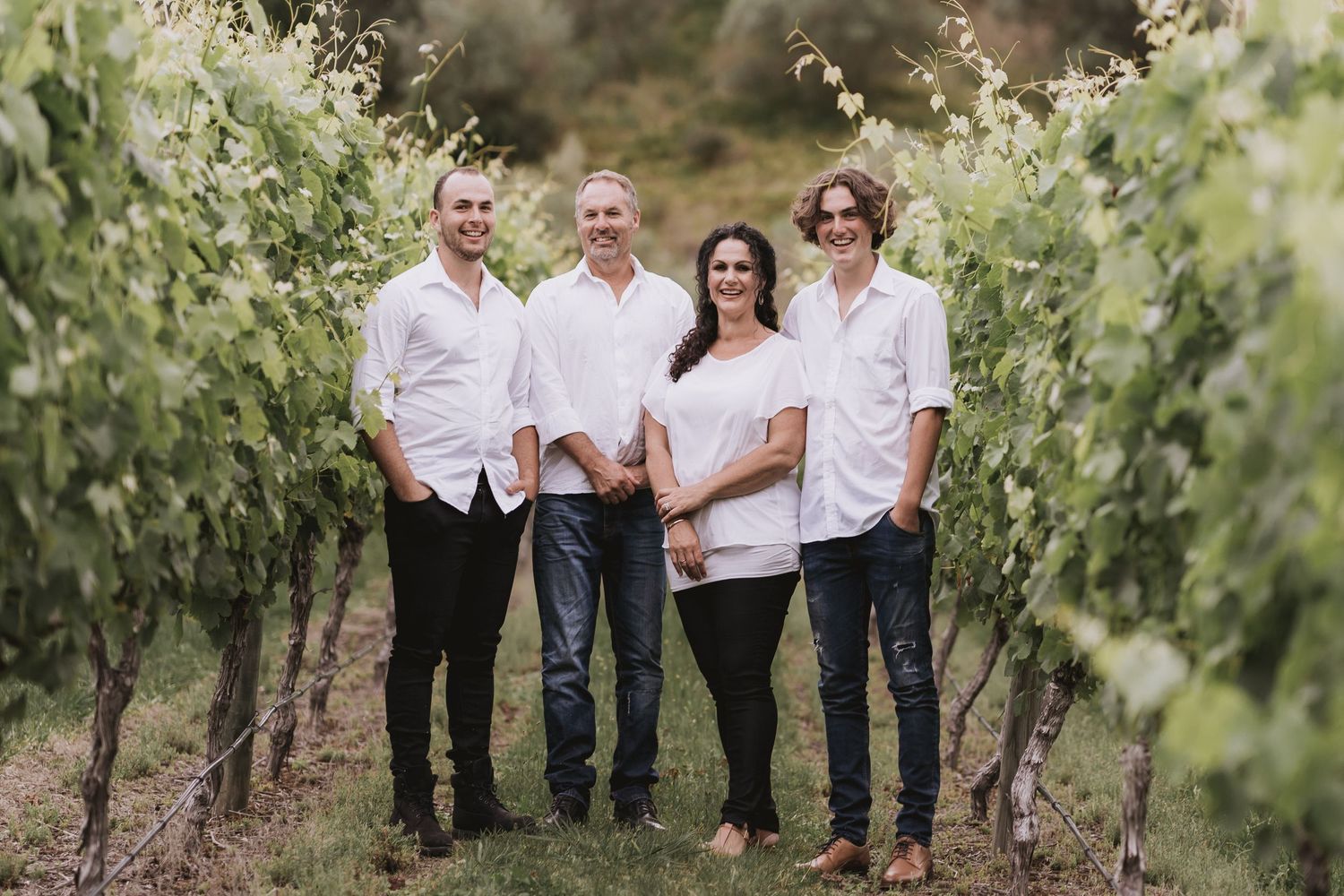 Family poses together in a casual outdoor vineyard setting wearing coordinating white shirts and jeans in natural light.