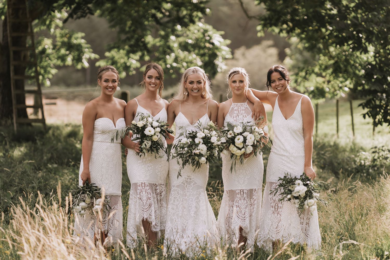 A group in white bridal party dresses stands in a sunlit meadow holding elegant floral bouquets.
