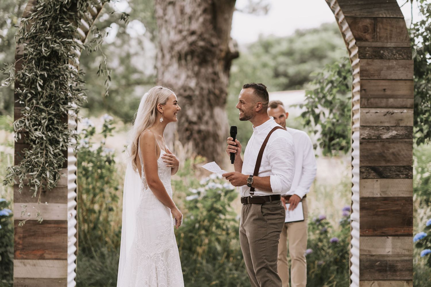 Under a wooden archway adorned with greenery, a groom delivers heartfelt wedding vows while the bride looks on with joy.