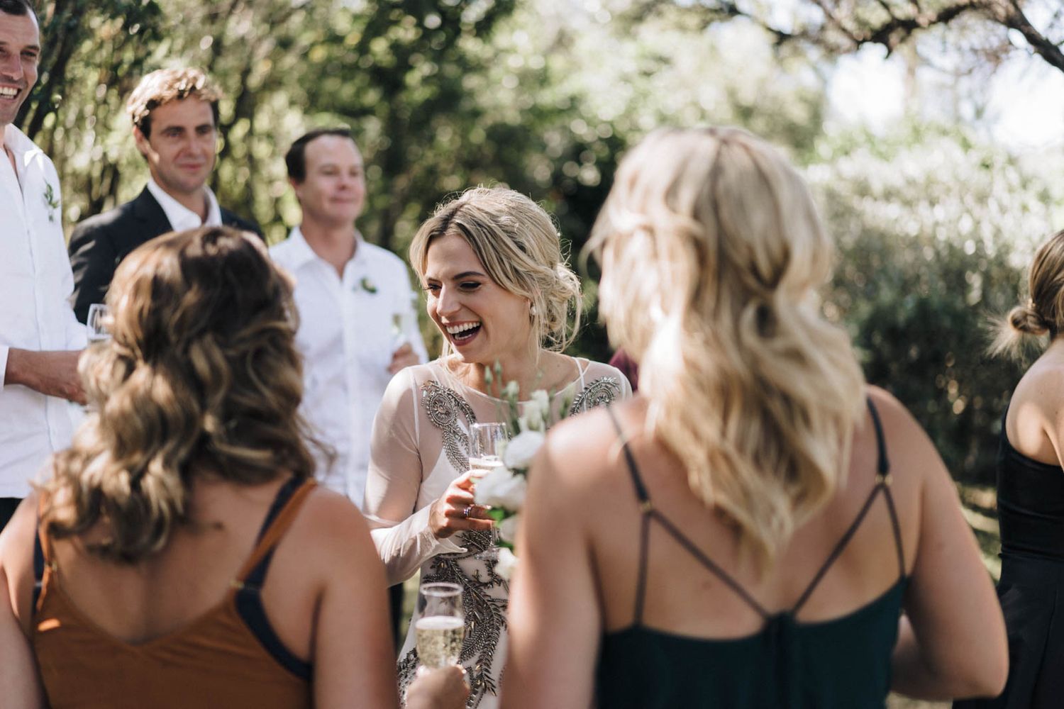 Wedding guests enjoying drinks and laughter during an outdoor celebration in natural sunlight.