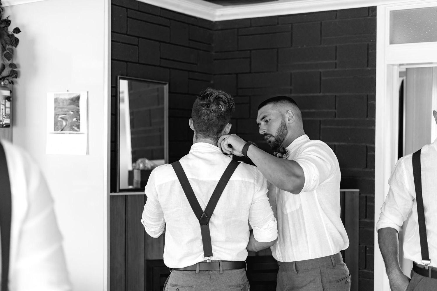 Groomsmen help with adjusting formal attire while getting ready for a wedding celebration.