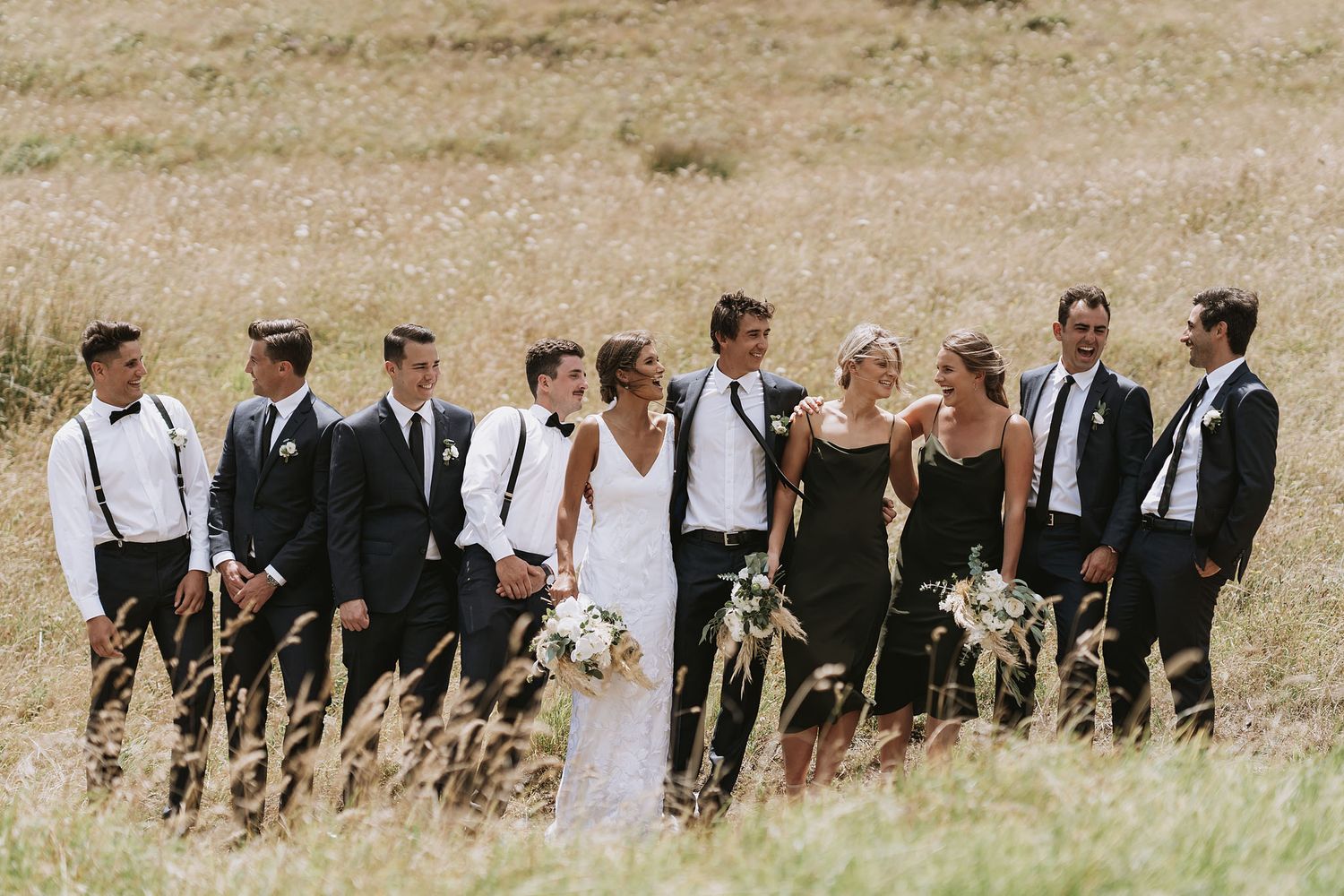 Wedding party in formal attire walking through a scenic grassy field on a bright summer day.