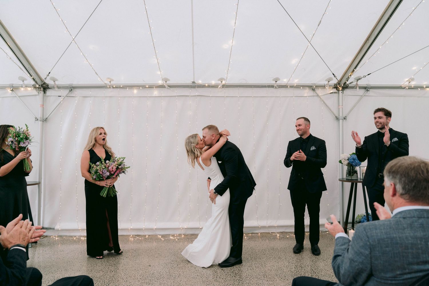 A couple shares their first kiss as newlyweds under a white tent while guests look on and applaud.