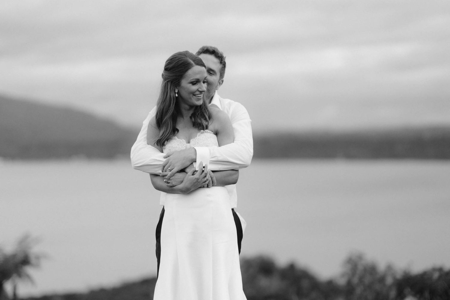 A black and white romantic portrait of a couple embracing by a lake with mountains in the background.
