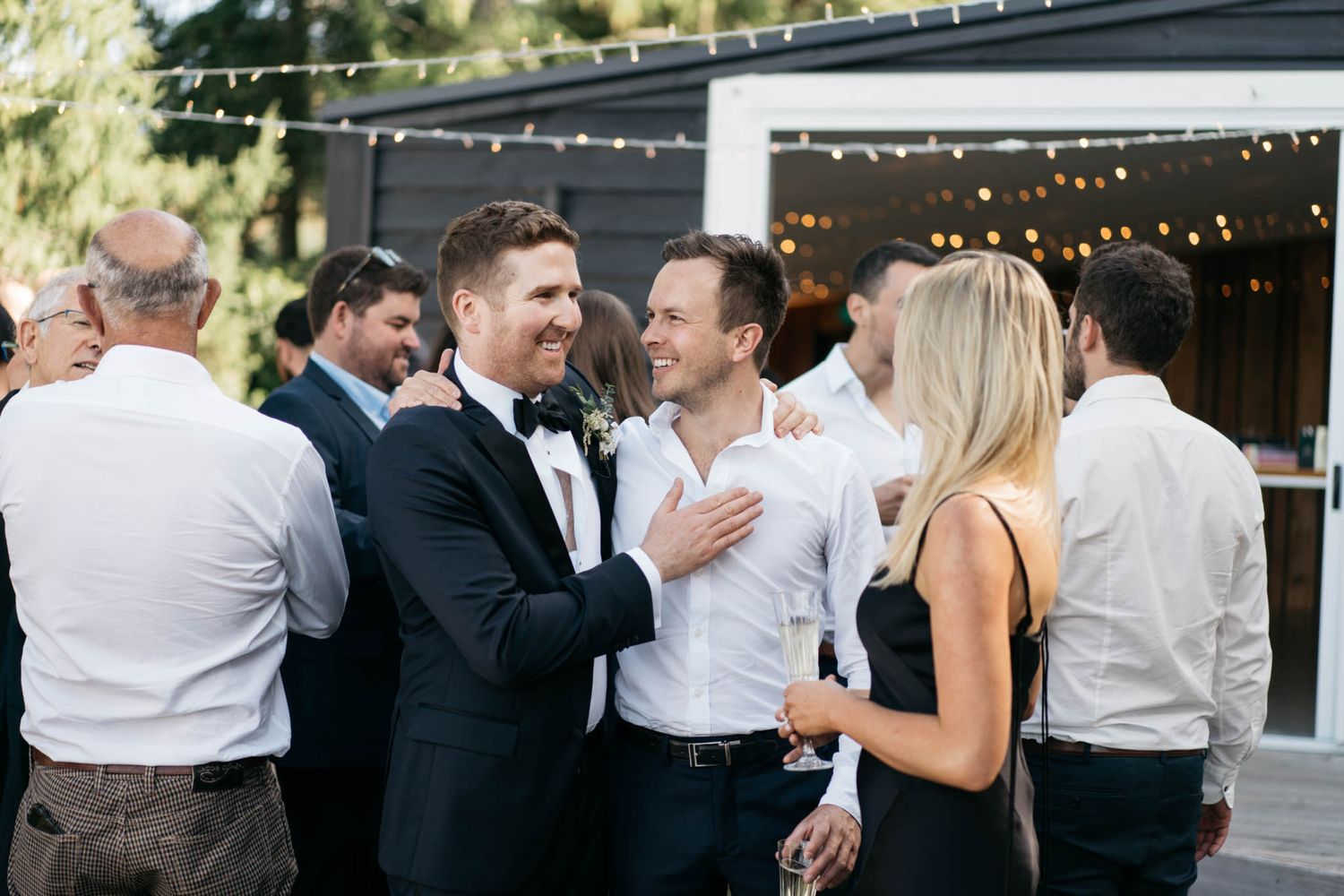 A group of wedding guests mingle and socialize on an outdoor patio decorated with string lights at a reception.