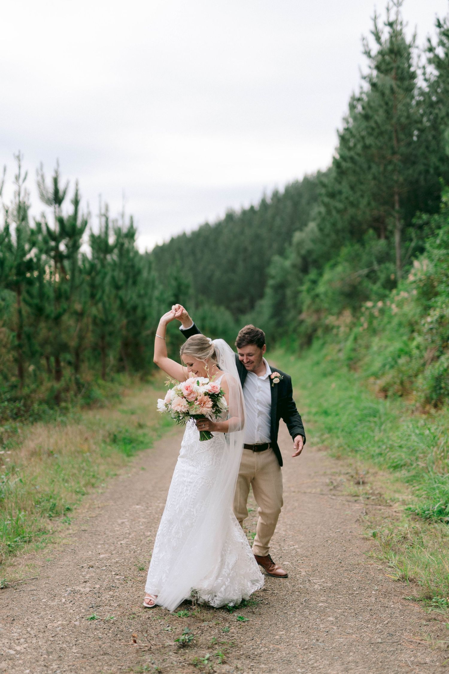 A couple celebrates and dances joyfully on a dirt path surrounded by lush evergreen forest on their wedding day.
