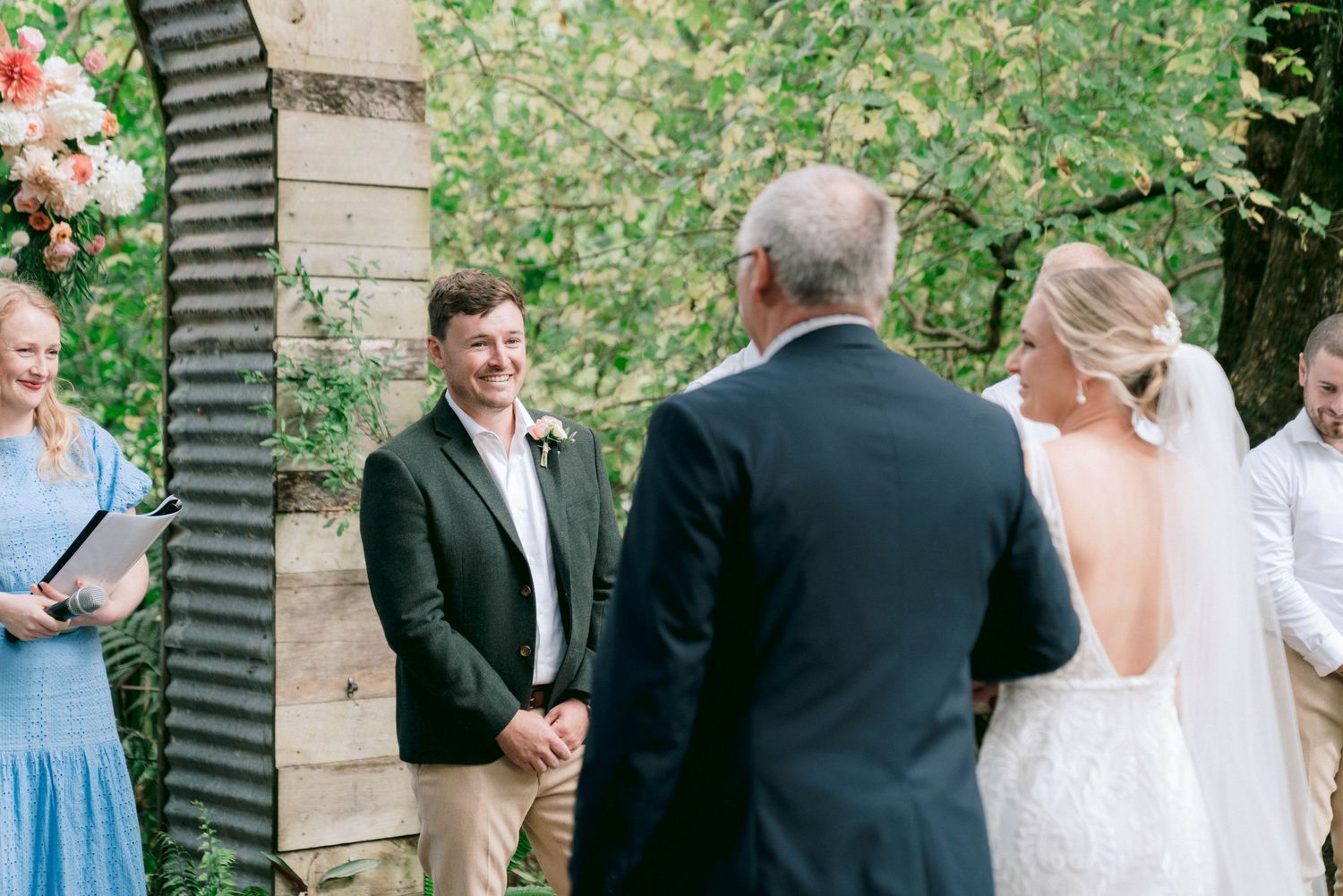 Traditional wedding ceremony taking place under decorative garden arch with pink flowers.