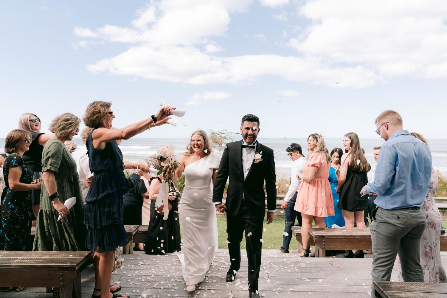 Newlyweds walk down the aisle while guests celebrate and cheer at an outdoor ceremony.