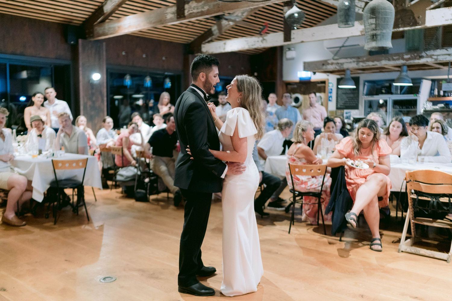 Dance floor moment during indoor wedding reception with guests seated at tables in background.