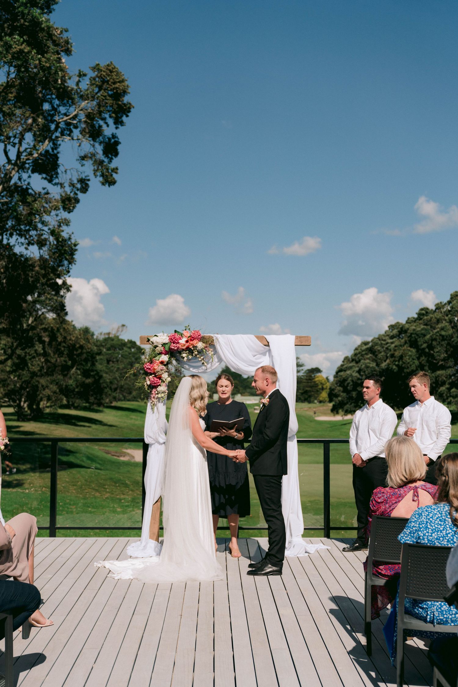 An outdoor wedding ceremony takes place on a wooden deck with a white arch and floral decorations against a scenic golf course.