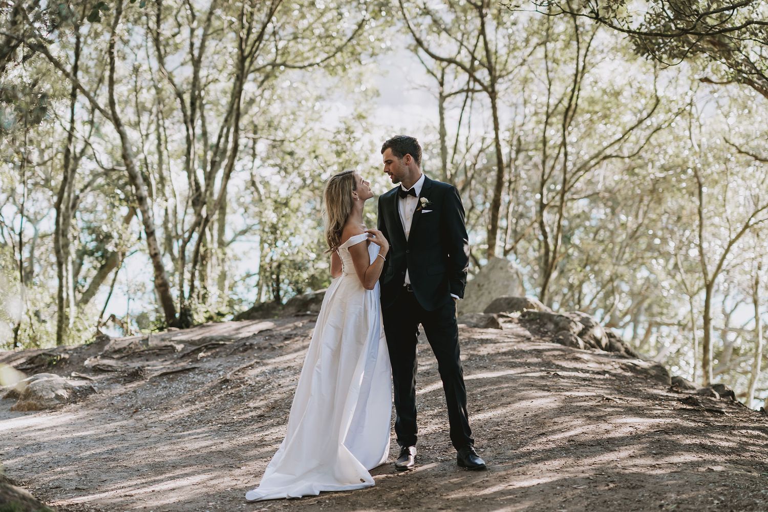 A couple in wedding attire walk together on a sunlit forest path surrounded by tall trees on their special day.