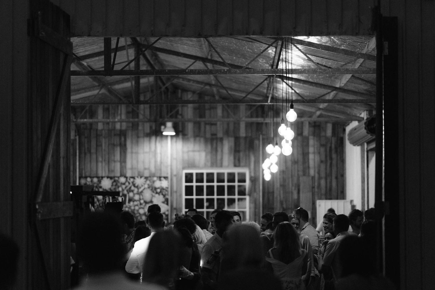 A dimly lit rustic venue with hanging pendant lights and guests gathered for an evening event in black and white.