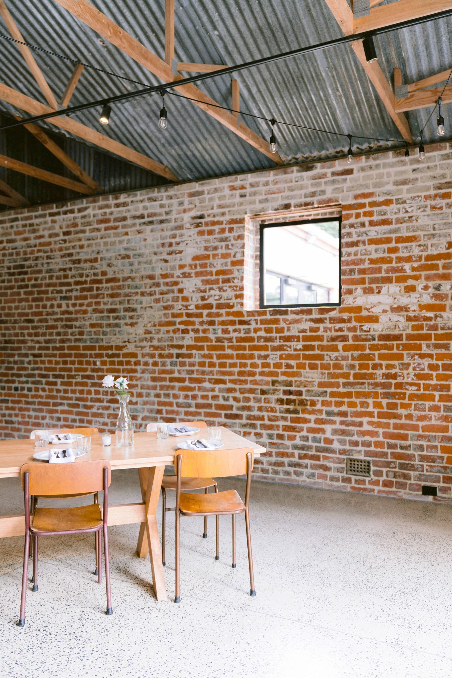 Rustic venue interior with exposed brick walls, wooden tables and chairs under metal roof beams.