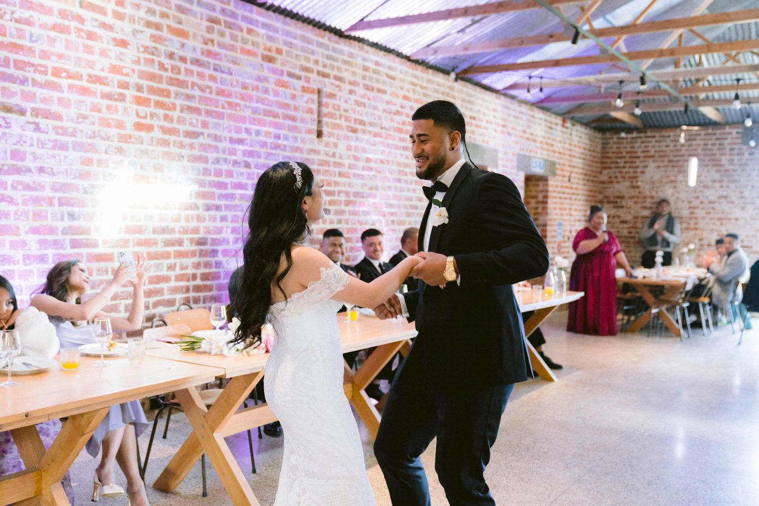 Couple dancing together in an industrial-style reception venue with exposed brick walls and wooden tables.