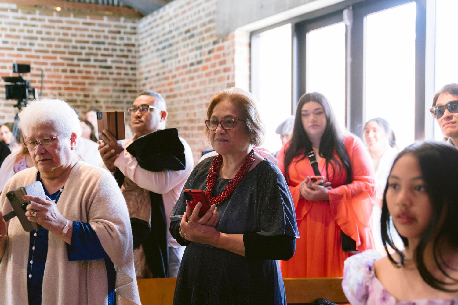 Wedding guests holding phones to capture moments inside brick-walled ceremony space.