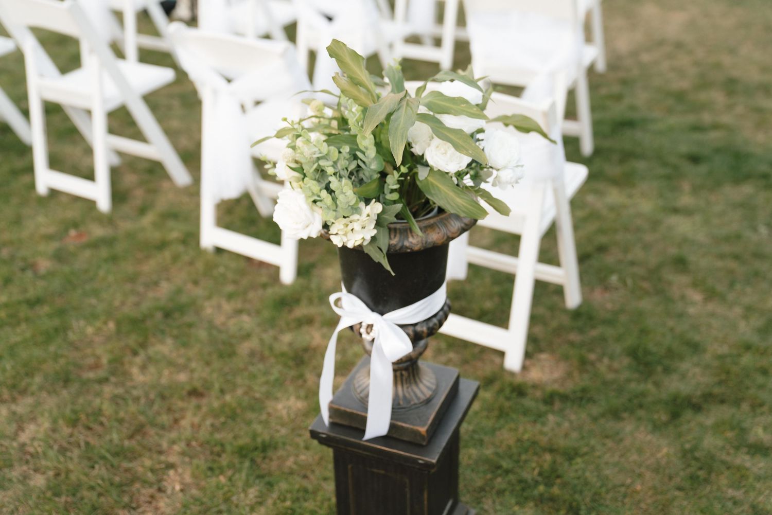 White folding chairs decorated with floral arrangements set up for outdoor wedding ceremony.