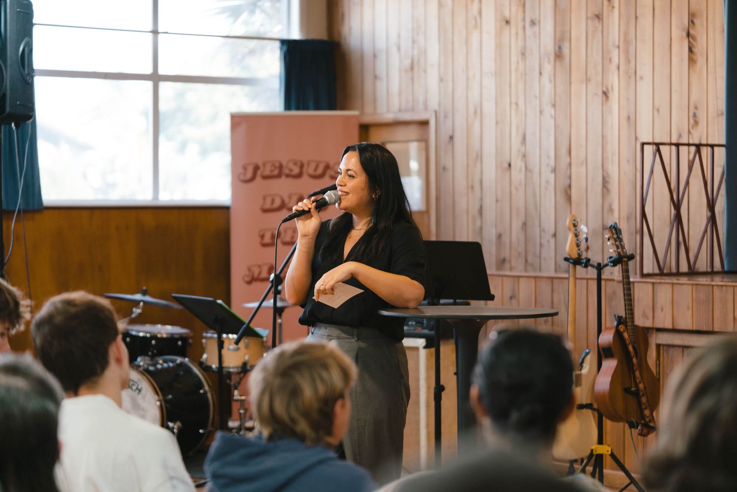 A worship leader singing into a microphone on stage with wooden paneled walls.