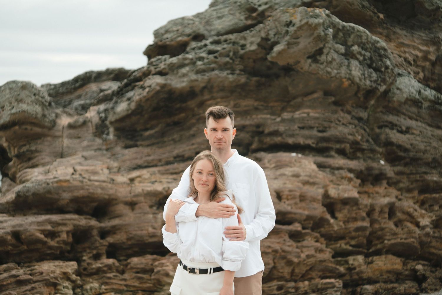 A couple in white outfits embraces while standing against rugged coastal rocks on an overcast day.